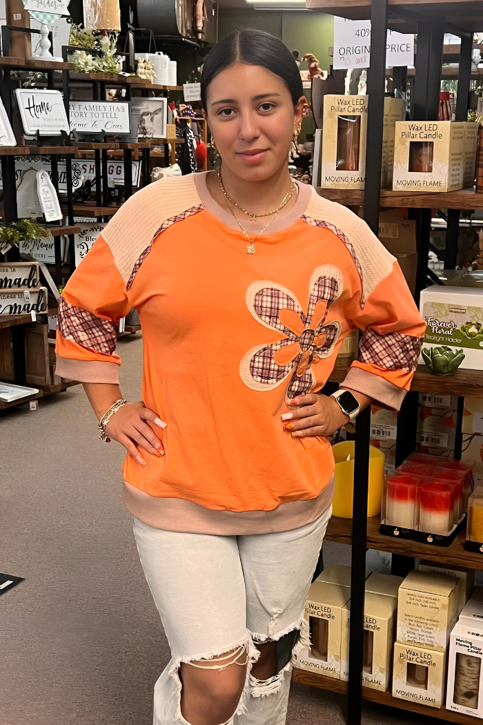 Wearing an orange long-sleeved shirt with patterned sleeves and a large floral appliqué on the front, and light-colored ripped jeans.