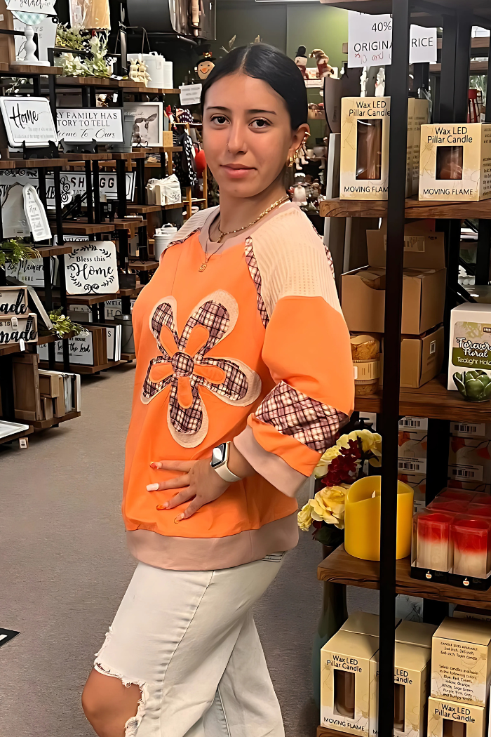 A woman wearing an orange long-sleeved shirt with a large plaid flower design on the front stands in what appears to be a retail store, looking towards the viewer.