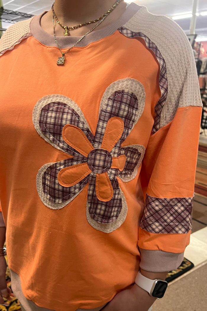 A close-up of an orange long-sleeved shirt with a large, plaid fabric flower applique on the front and plaid accents on the sleeves and collar. 