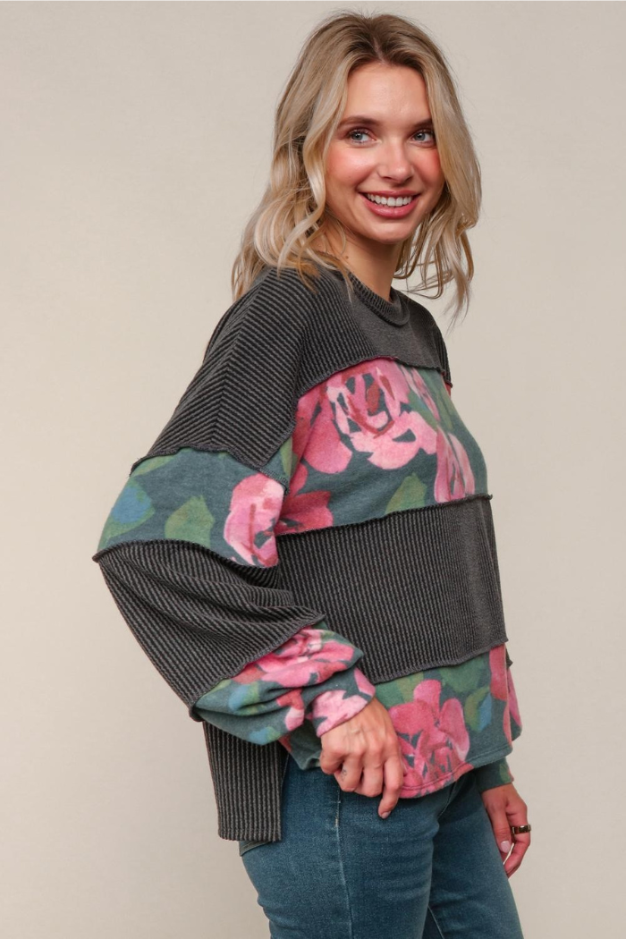 A blonde woman smiling and looking slightly off-camera to her right, wearing a long-sleeved, loose-fitting top with a patchwork design. The top features alternating horizontal stripes of dark gray ribbed fabric and sections with a floral pattern of pink roses on a dark green background.