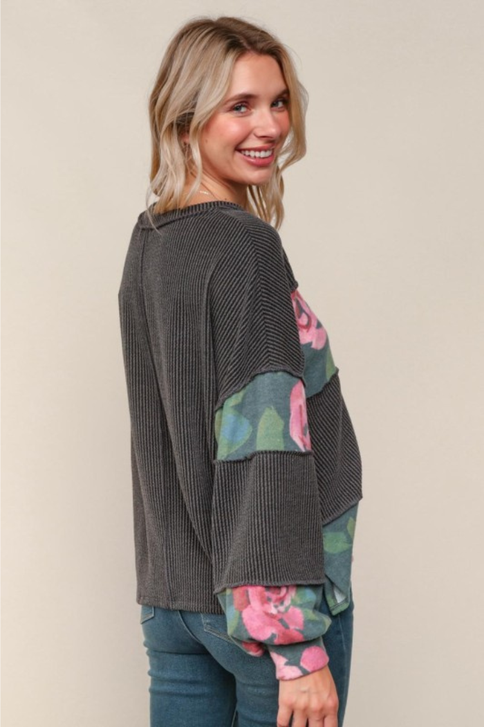A woman with blonde hair smiles while looking over her right shoulder. She is wearing a dark gray ribbed long-sleeved top with floral print panels in shades of pink, green, and blue on the sleeves and sides. She is also wearing blue jeans. The background is a plain light tan color.
