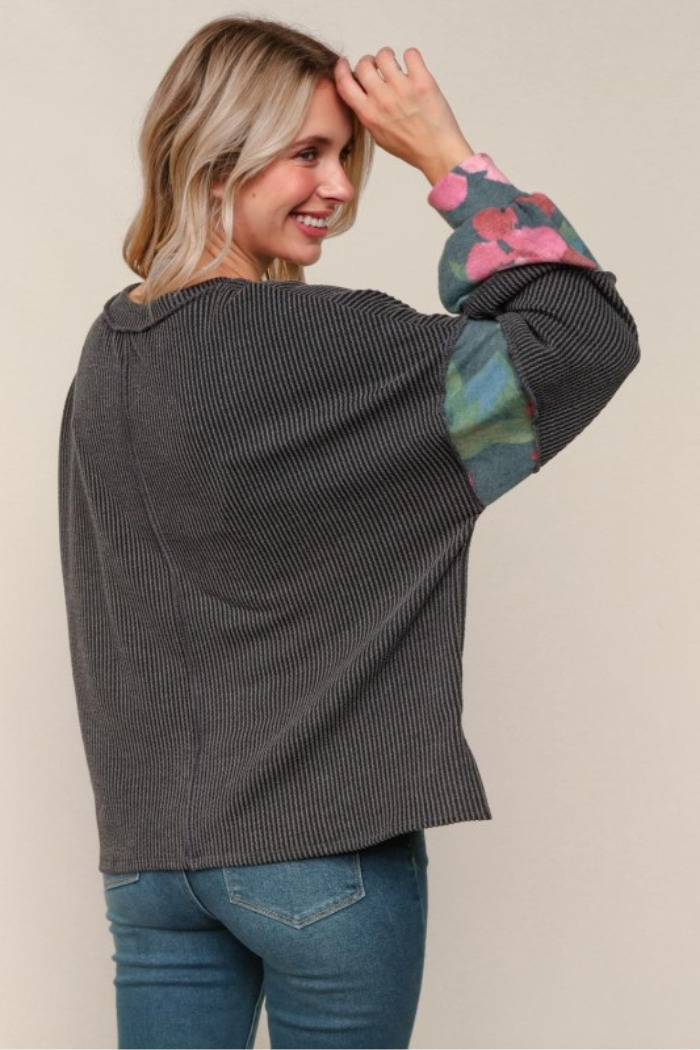 A woman with blonde hair, facing away and looking over her shoulder with a smile, wears a dark grey ribbed long-sleeved top with colorful floral patterns on the sleeves, paired with blue jeans.