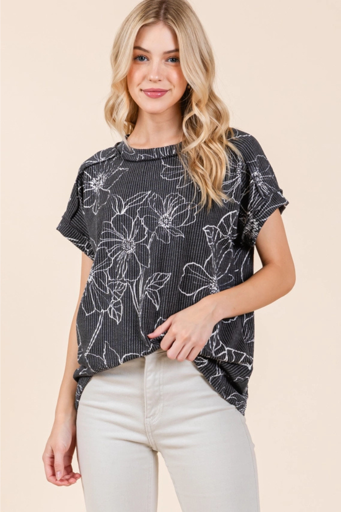 A woman with blonde hair and a relaxed expression models a charcoal grey, short-sleeved, ribbed knit tee with a white floral print, paired with light-colored pants.