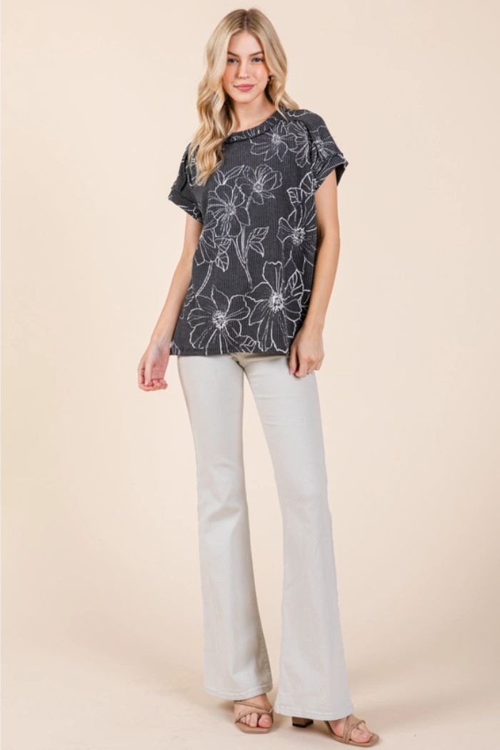 A young woman stands facing forward. She has long, wavy blonde hair and is wearing a dark gray, short-sleeved, relaxed-fit top with a subtle rib-knit texture and an abstract white floral pattern.