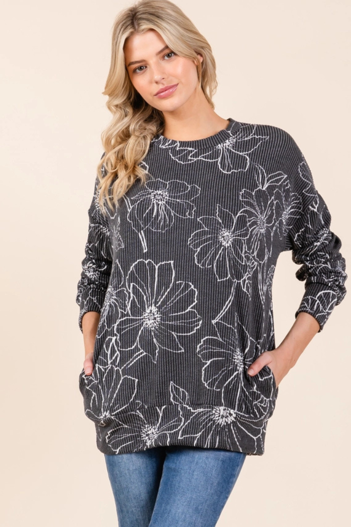 A young woman with blonde hair stands, looking forward and slightly smiling, wearing a charcoal long-sleeve top with a prominent white floral outline pattern, textured with a ribbed fabric. She has her hands casually placed in the front pockets of the top, which extends down past her hips, and is paired with blue jeans. The background is a plain, light color.