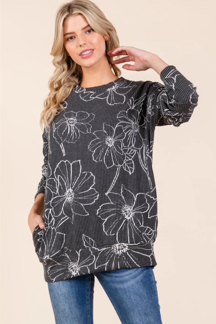 A blonde woman smiling and looking at the camera, wearing a dark charcoal gray, ribbed long-sleeved top with large, white floral outlines all over it. She has her left hand in her pocket and her right hand near her hair.