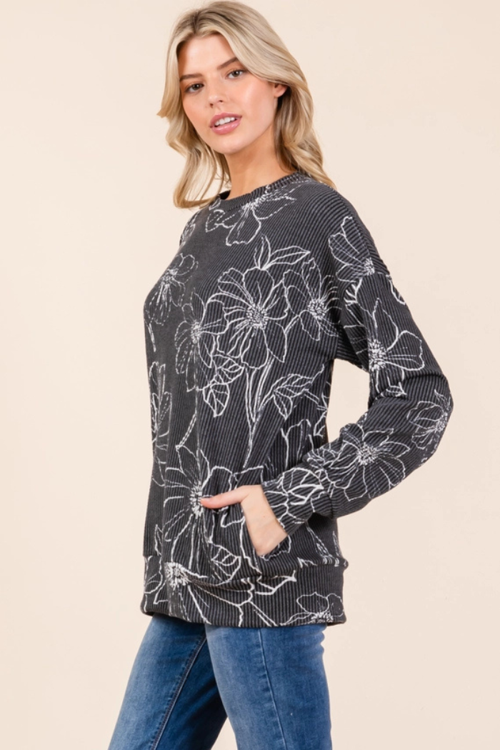 A woman wearing a dark grey, ribbed knit, long-sleeved top featuring a white floral pattern and front pockets, paired with blue jeans. She stands with one hand in a pocket.