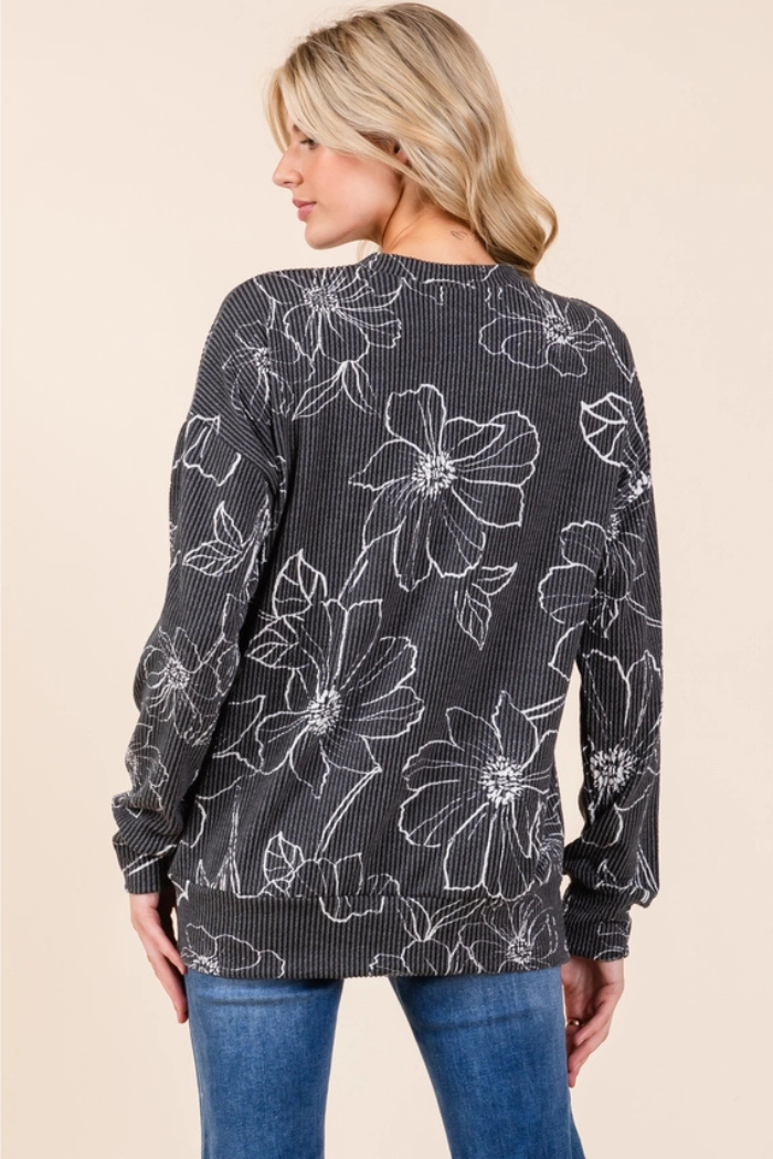 A woman seen from the back, wearing a dark grey ribbed long-sleeved top with white outline floral patterns, paired with blue jeans.