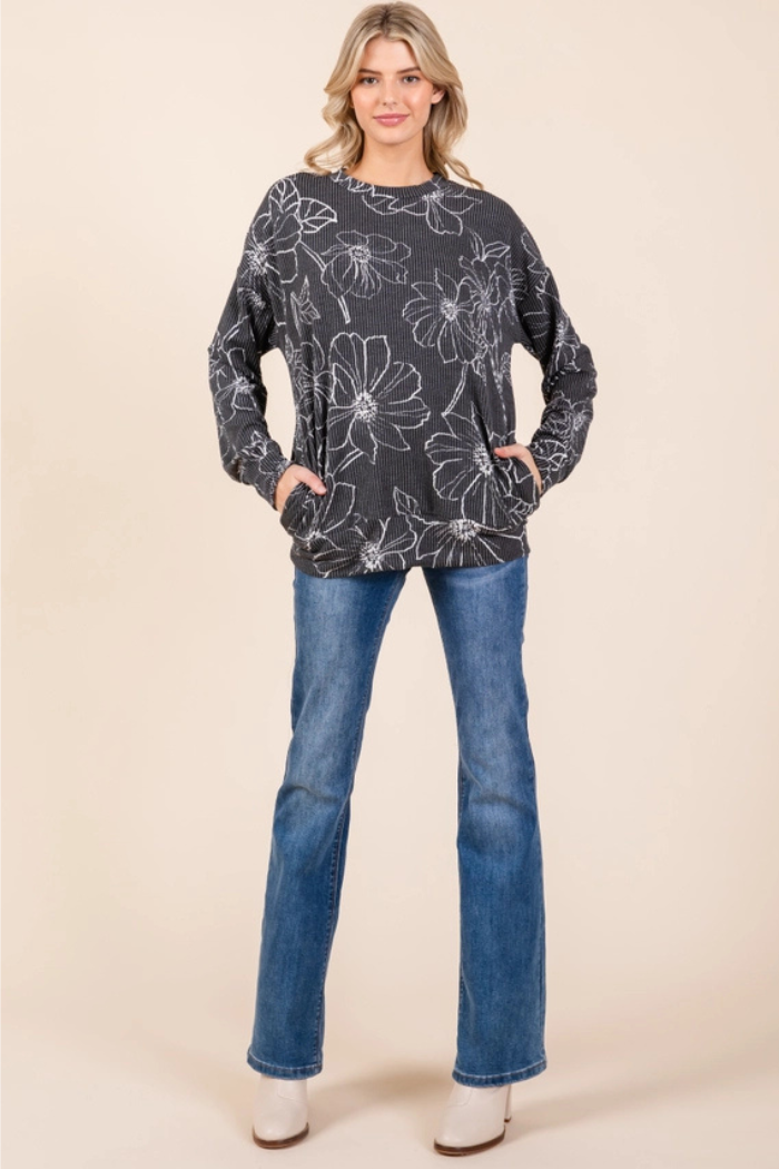 A woman wearing a charcoal grey long-sleeved top with a white floral outline pattern and blue jeans with white boots. The top has pockets on the front.