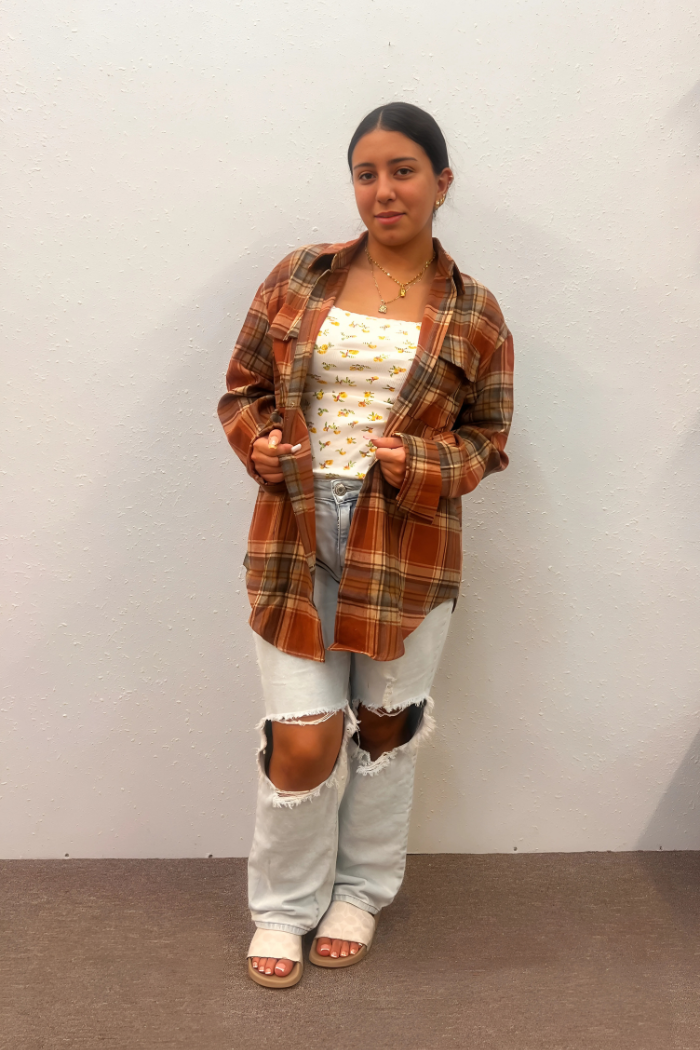 A young woman with dark hair pulled back, wearing a brown and orange plaid oversized shirt unbuttoned over a white floral print top, light-wash ripped jeans, and light-colored sandals, stands facing forward against a plain light-colored wall.