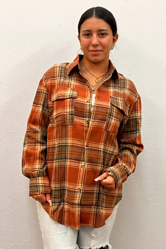 A young woman with dark hair stands facing forward, wearing a loose-fitting, long-sleeved flannel shirt with a brown, orange, and beige plaid pattern.