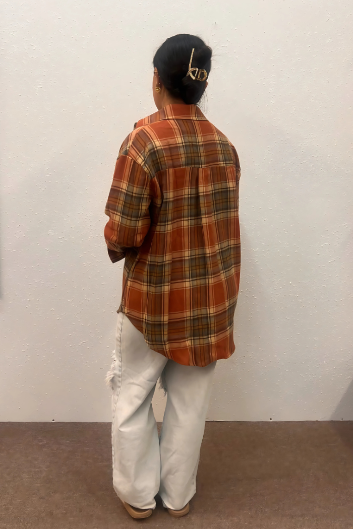 A woman seen from the back wears an oversized, loose-fitting plaid shirt in shades of orange, brown, and green, paired with light-wash ripped jeans. The shirt features a drop shoulder design and appears to be made of a soft, flannel-like material. 