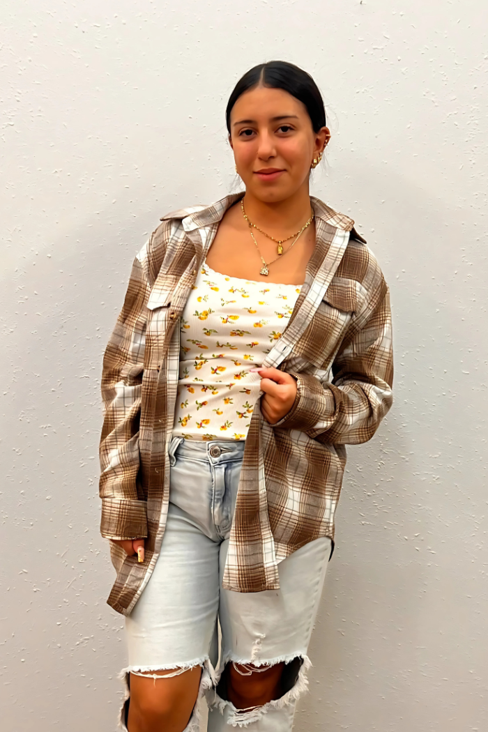 A young woman with dark hair stands facing forward, wearing a brown and white plaid flannel shirt unbuttoned over a white tank top with a yellow floral print. She is also wearing light-wash ripped jeans and a gold necklace with multiple charms.