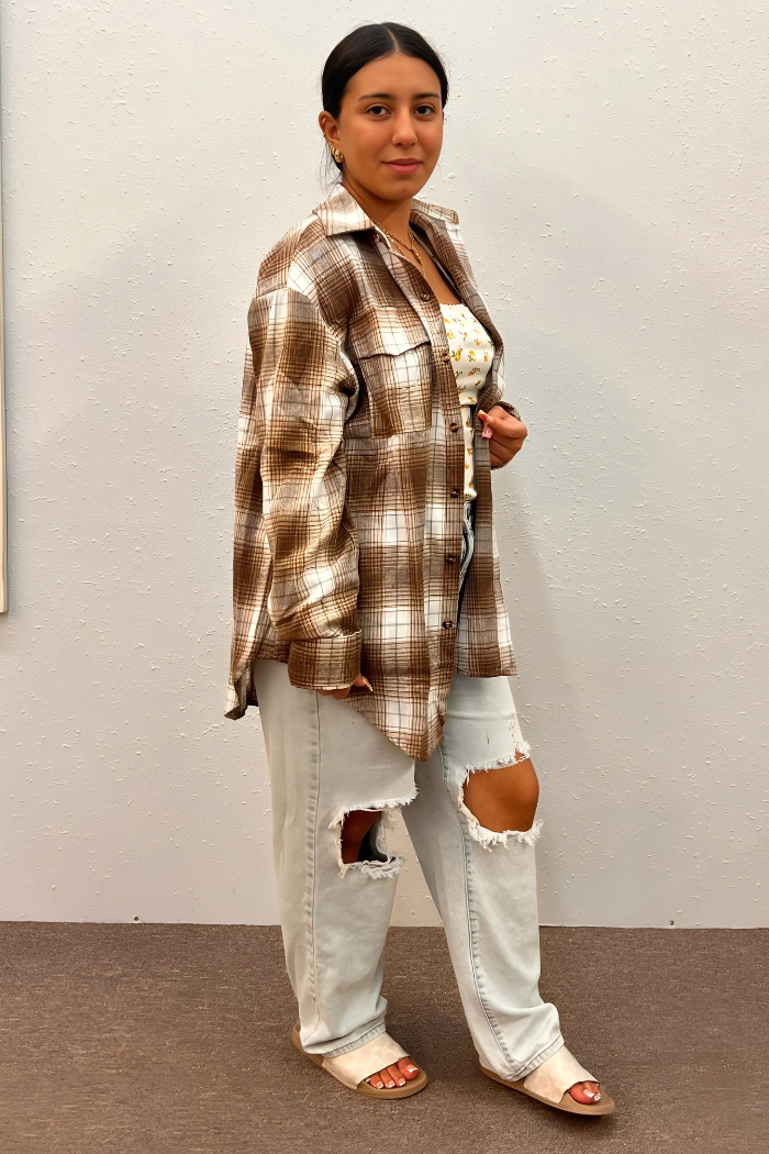 A woman wearing a brown and white plaid flannel shirt over a white floral top, light-wash ripped jeans, and light-colored slide sandals, standing against a light gray wall.
