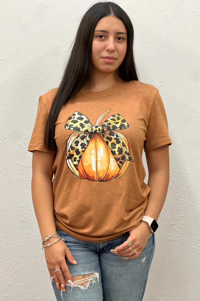 A woman stands facing forward, wearing a short-sleeved, brownish-orange t-shirt featuring a graphic of an orange pumpkin adorned with a leopard print bow. She is also wearing blue jeans and has multiple bracelets on her wrist and rings on her fingers.
