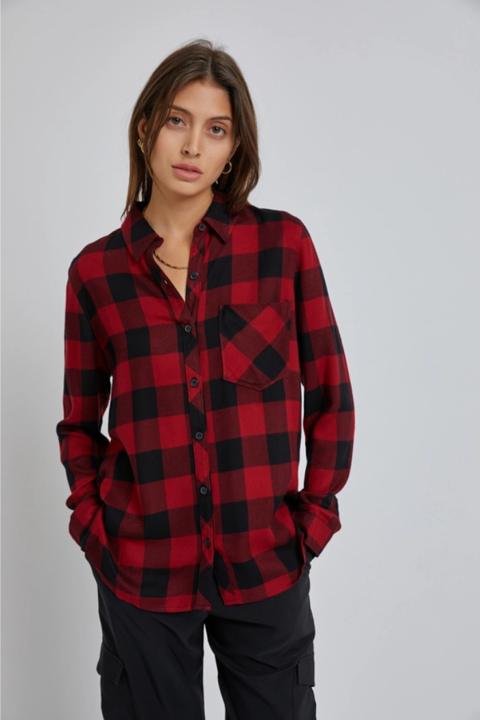 A woman wearing a red and black plaid flannel shirt, black cargo pants, and a gold chain necklace, stands with her hands in her pockets, looking directly at the viewer. 