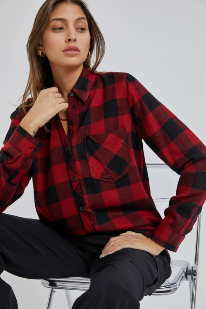 A woman wearing a red and black plaid flannel shirt and dark pants is seated, looking towards the viewer. The shirt features a button-down front and a patch pocket on the left side of the chest. She has long, dark hair, and is wearing hoop earrings. 