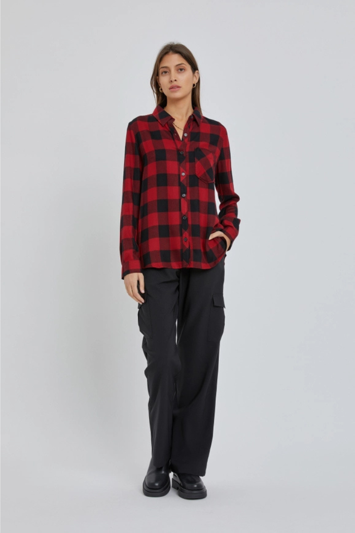 A full body shot of a woman standing and facing forward, wearing a red and black plaid flannel shirt with long sleeves and black cargo pants. She is also wearing black shoes. Her left hand is in her pocket.