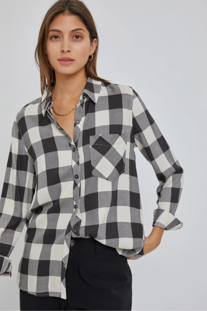 A woman wearing a red and black plaid flannel shirt, black cargo pants, and a gold chain necklace, stands with her hands in her pockets, looking directly at the viewer. 