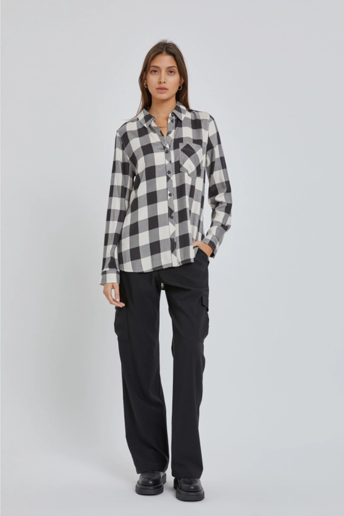 A full-body shot of a woman standing, wearing a black and white plaid long-sleeve button-down shirt and black wide-leg cargo pants. She has her left hand in her pocket and is looking towards the camera.