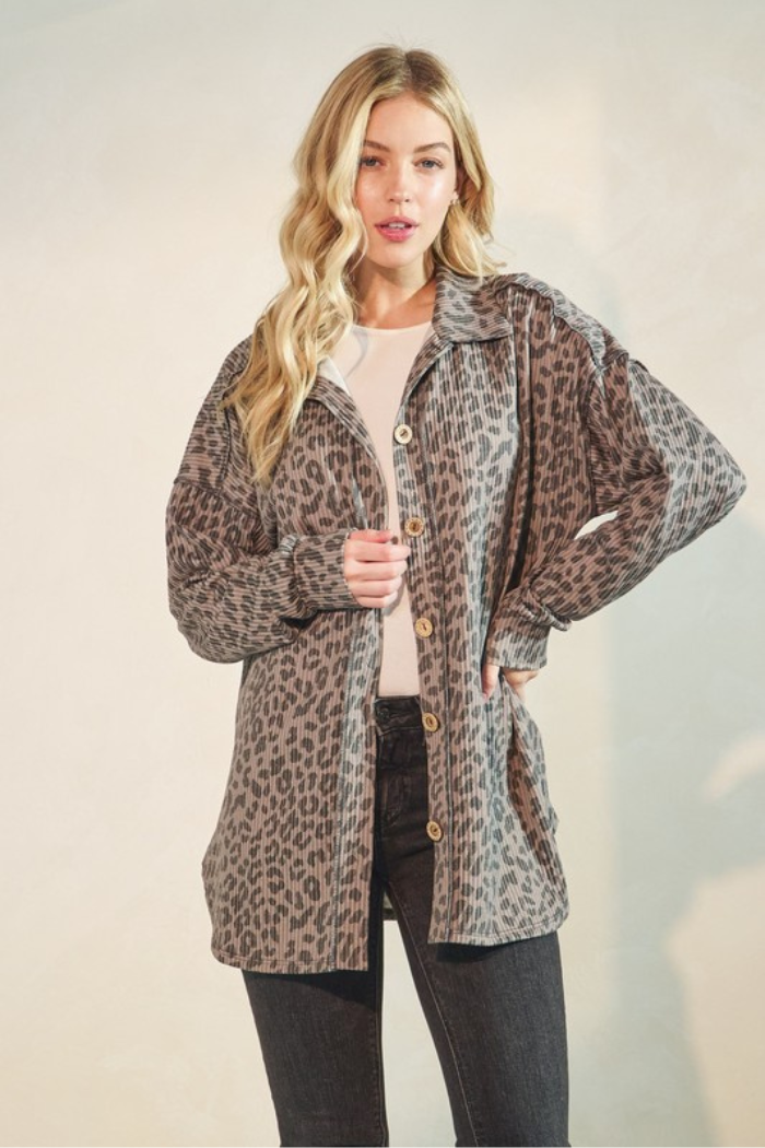 A blonde woman wearing a taupe and black animal print button-up over a white top and dark jeans, standing against a cream-colored background. 
