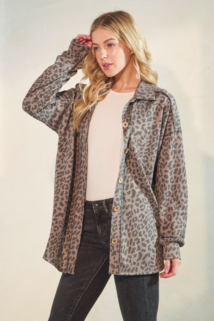 A woman with blonde hair wearing a taupe and black animal print long sleeve top over a white top and black pants. She stands facing forward with one hand near her hair.