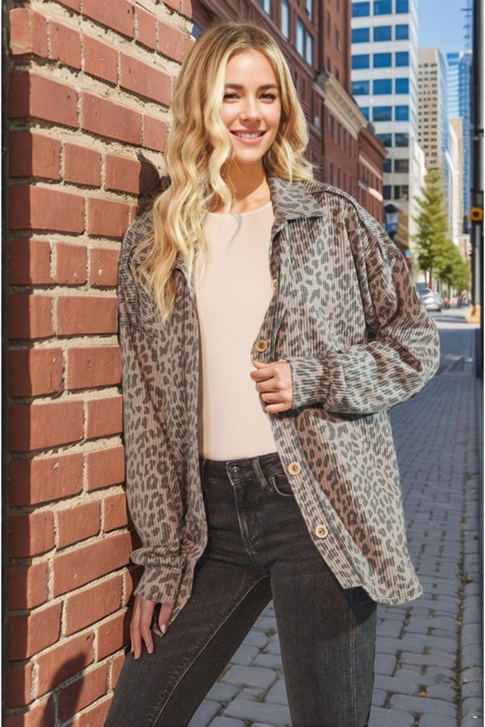 A young woman with long blonde hair smiles at the camera, leaning casually against a brick building on an urban street. She is wearing a taupe and black animal print long sleeve over a light-colored top, paired with dark wash jeans. In the background, there's a paved street and modern buildings under a clear sky, suggesting a city setting.