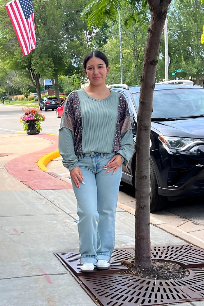 A young woman with dark hair stands on a sidewalk next to a tree, wearing a ribbed olive, long-sleeved top with patterned accents on the shoulders and light-wash jeans. 