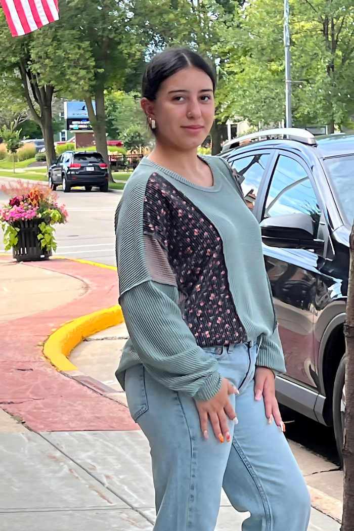 A young woman with dark hair pulled back stands outdoors next to a black car and a sidewalk planter with flowers. She is wearing an Olive-colored long-sleeved top with a floral-patterned section and light-wash jeans. She is looking towards the camera and smiling slightly.