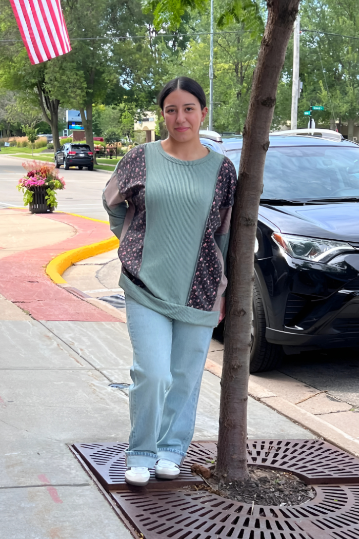 Woman standing on a sidewalk next to a tree, with a car and street in the background. She is wearing an Olive-colored long-sleeved top with a floral-patterned section, oversized and light-wash jeans. 