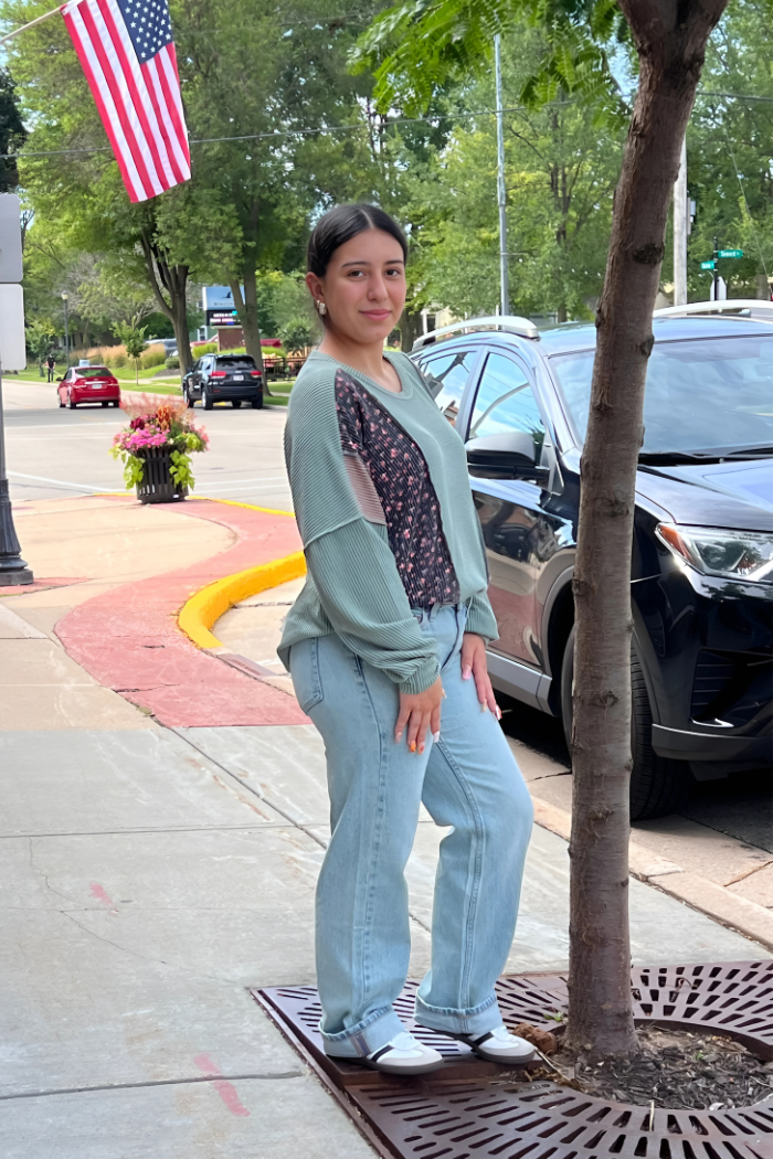 A young woman is wearing an Olive-colored long-sleeved top with a floral-patterned section and light-wash jeans. She is looking towards the camera and smiling slightly. Standing from the side.