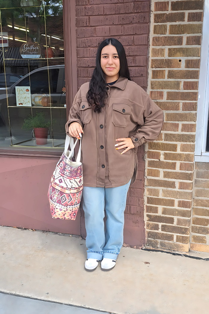 A young woman with long, dark hair stands smiling in front of a brick building and a storefront window, holding Southwestern or Aztec print canvas tote. She is wearing a brown shacket (shirt-jacket), light blue wide-leg jeans, and white sandals.
