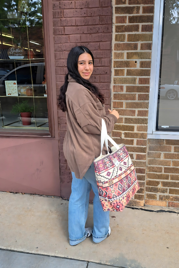 A young woman with dark, wavy hair stands facing slightly away from the camera, looking back over her shoulder with a smile. She is wearing a brown long-sleeved shacket and light blue jeans. She carries a large, Southwestern or Aztec print canvas tote on her left shoulder. 