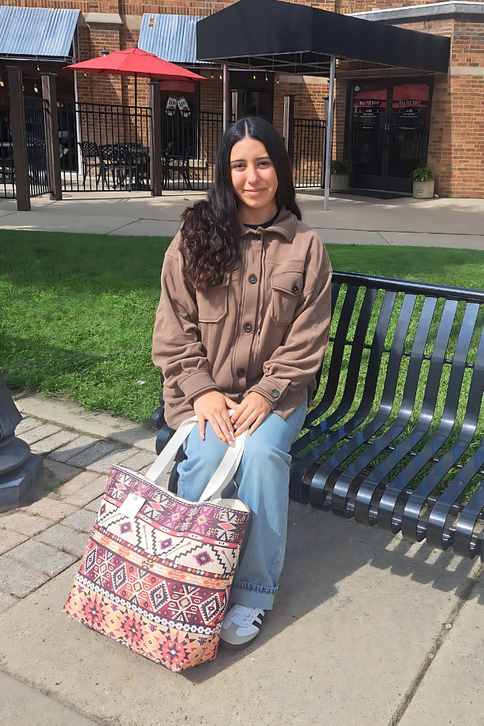 A young woman with dark hair sits on a black slatted bench outdoors, holding a large, Southwestern or Aztec print canvas tote. She is wearing light-wash jeans, white sneakers with black stripes, and a loose-fitting, button-up brown shacket. 