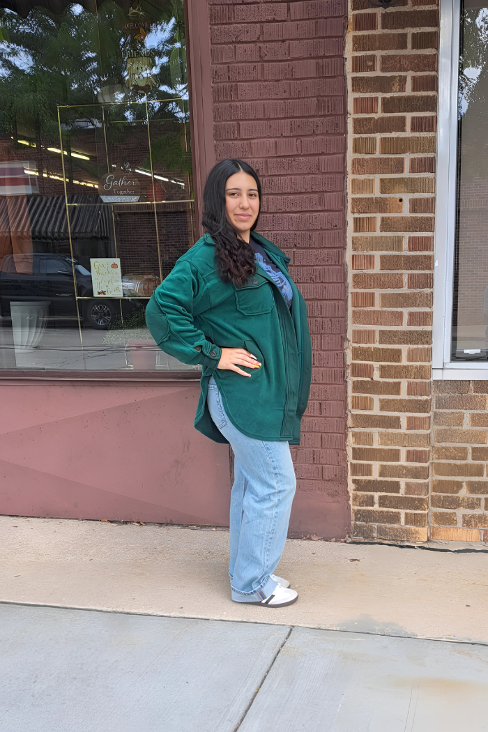 A woman with long, dark, curly hair poses on a sidewalk in front of a brick building, wearing a dark green oversized shacket, light-wash jeans, and white sneakers.