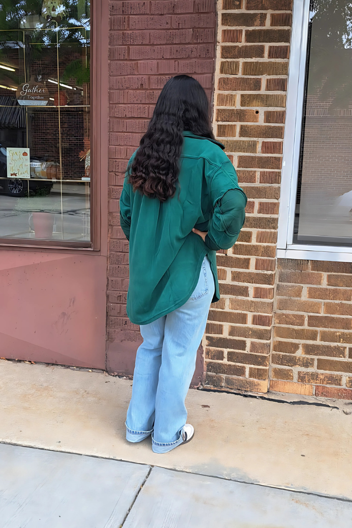This image shows a person from behind, with long, dark, wavy hair, standing on a sidewalk in front of a brick building. They are wearing a loose-fitting, oversized green shacket, light blue jeans, and light-colored shoes with a dark accent. 