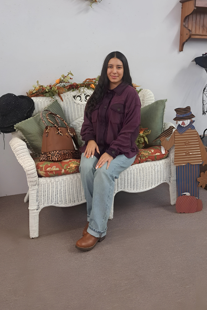 A young woman with long, dark hair sits on a white wicker bench adorned with green and orange pillows and fall-themed decor. She is wearing a dark purple shacket, light-wash blue jeans, and brown shoes. A brown and leopard-print handbag rests on the bench next to her.