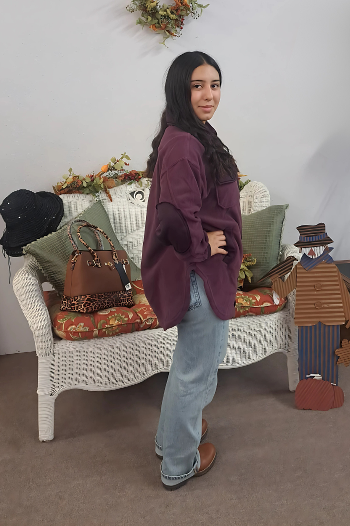 A young woman with long dark hair, wearing an oversized dark purple shacket with elbow patches, light-wash jeans, and brown shoes, stands facing slightly right, looking towards the viewer with a slight smile. She is positioned to the right of a white wicker bench decorated with fall-themed items like pumpkins, leaves, and a scarecrow figure. A brown purse with a leopard print strap rests on the bench next to a black hat. The background appears to be a light-colored wall.
