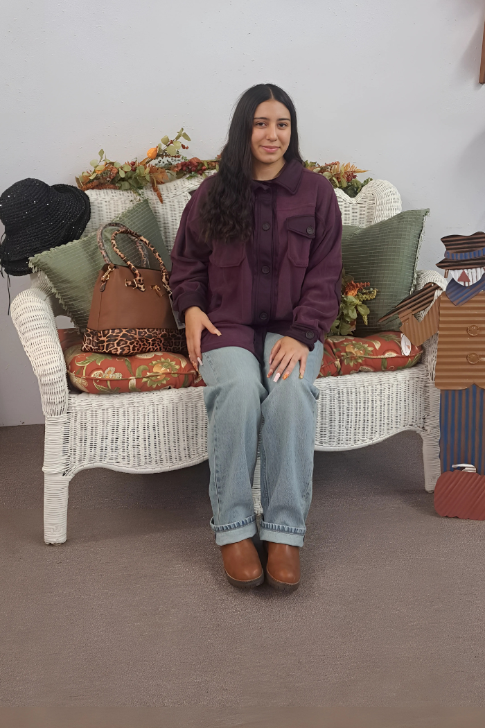 A young woman with dark, long hair sits centered on a white wicker bench, smiling slightly at the camera. She wears a dark purple shacket, light-wash blue jeans, and brown boots. A brown purse with a leopard print handle sits beside her on the bench. The bench is adorned with green pillows and autumn-themed decor, including garlands of leaves and berries. A black hat hangs on the left side of the bench, and a wooden scarecrow figure stands to the right, completing the cozy fall setting.