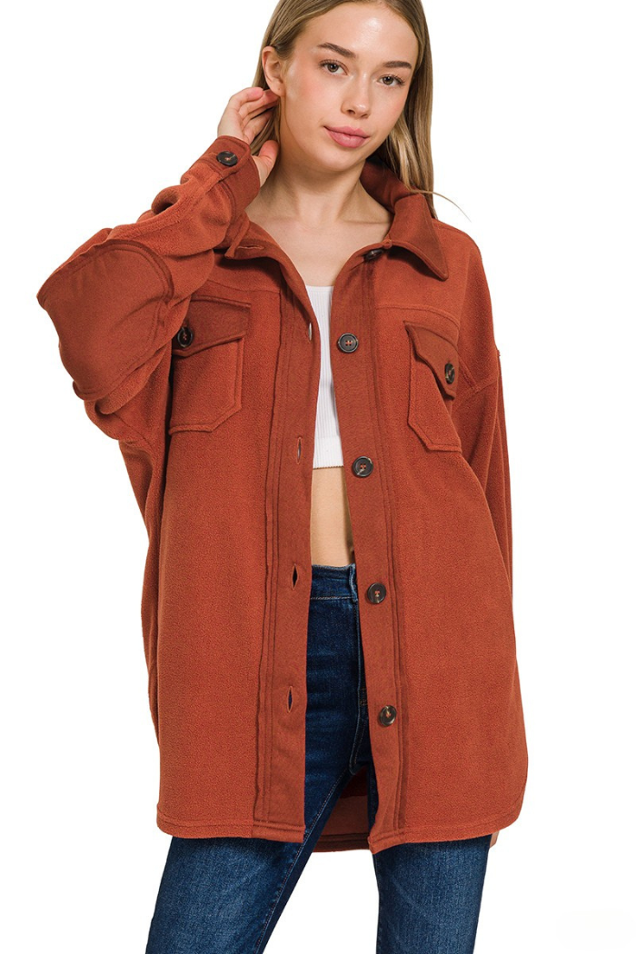A young woman with long blonde hair poses while wearing a rust-colored oversized fleece shacket over a white top and blue jeans, with a white background. The shacket features a button-front closure, collared neckline, and chest pockets with button flaps, made of a soft fleece material, and extends to about mid-thigh.