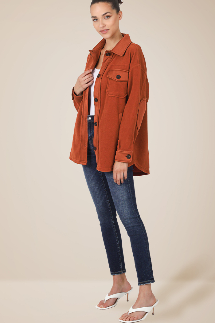 A woman with dark hair pulled back, wearing a rust-colored shacket over a white top, dark skinny jeans, and white high-heeled sandals. The shacket has large front pockets and button details, and she is standing with one hand holding the lapel of the jacket.