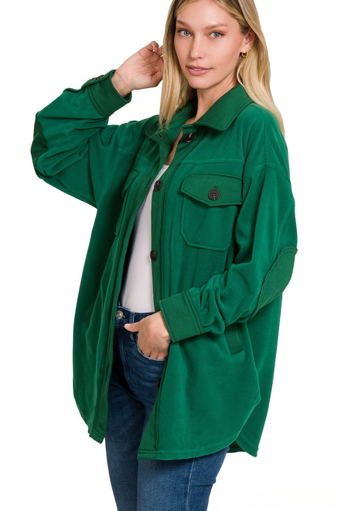 A blonde woman wearing a dark green oversized fleece shacket with a collared neckline, front button closure, two front flap pockets, button cuffs, and elbow patches, standing with one hand in her jeans pocket and the other near her head.
