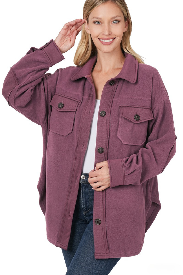 A woman wearing a dark plum-colored oversized fleece shacket with large buttons and front chest pockets, paired with blue jeans and a white top, stands against a white background. The shacket features a classic collar, dropped shoulders, and long sleeves with button cuffs, designed in a loose, relaxed silhouette. 