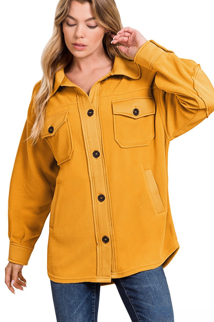 A woman with blonde hair wears an oversized mustard yellow fleece shacket with black buttons, chest pockets, and side pockets, paired with blue jeans. She stands against a white background, smiling slightly and looking forward. 