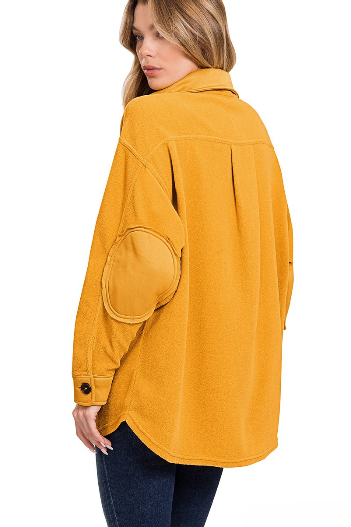 A woman from the back wears a mustard yellow, oversized shacket with a rounded patch pocket detail on the left elbow. The shacket appears to be made of a soft, possibly fleece-like material, and has a collared neckline and long sleeves with buttoned cuffs. The hem is curved. 