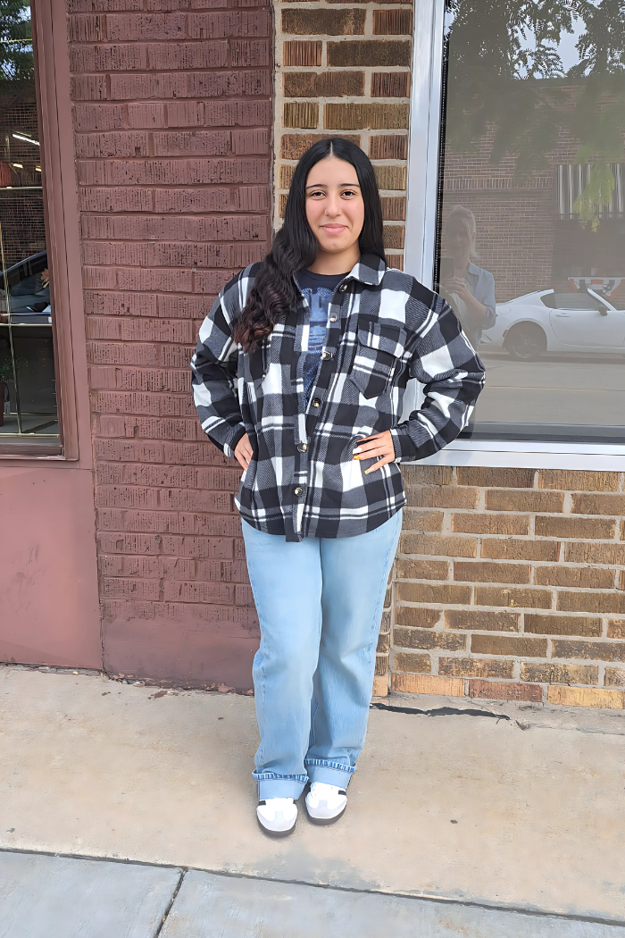 A young woman with long dark hair, wearing a black and white checkered fleece shacket over a dark t-shirt, light wash jeans, and white sandals, stands with hands in pockets outside a brick building with a large window reflecting a white car. 