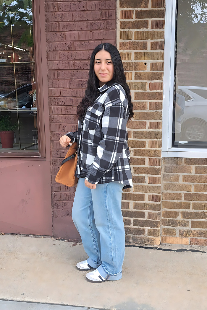 A young woman with long, dark hair stands smiling sideways in front of a brick building. She is wearing a black and white checkered fleece shacket, light-wash jeans, and white and black athletic shoes. She carries a brown tote bag on her left arm. 