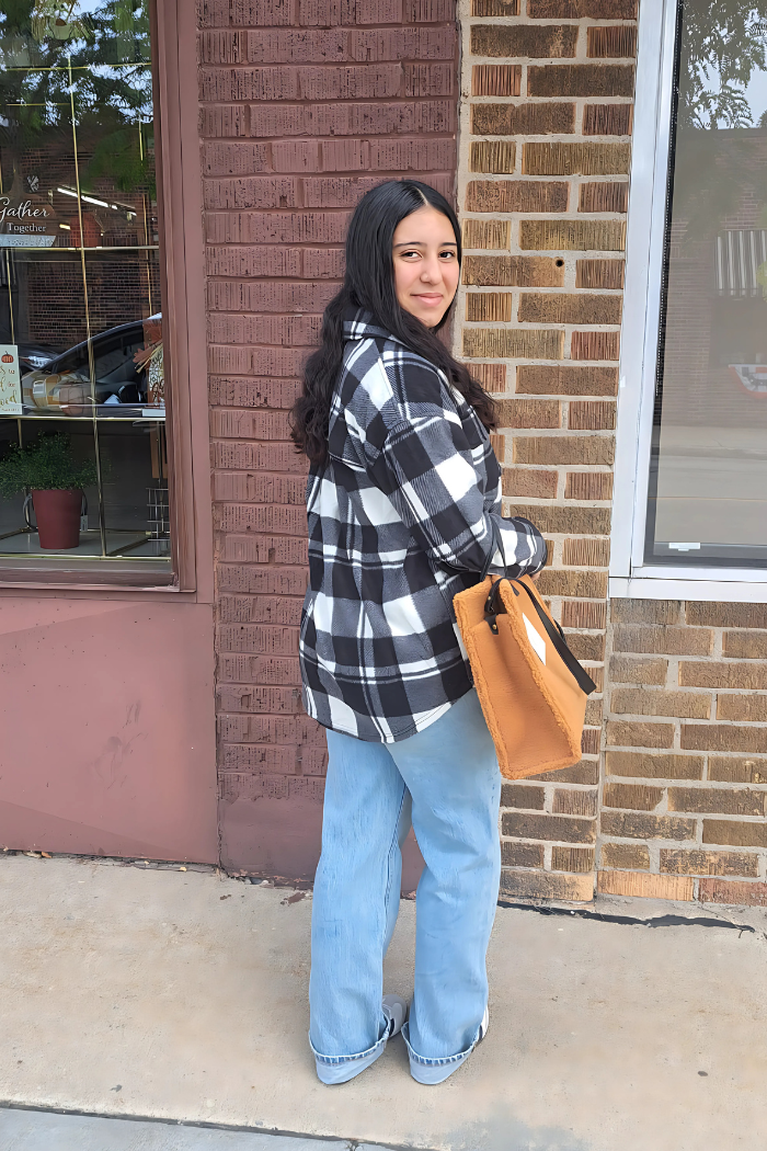 A young woman with dark hair, wearing a black and white checkered shacket, light blue jeans, and carrying a tan tote bag, stands smiling over her shoulder at the camera in front of a brick building with a shop window.