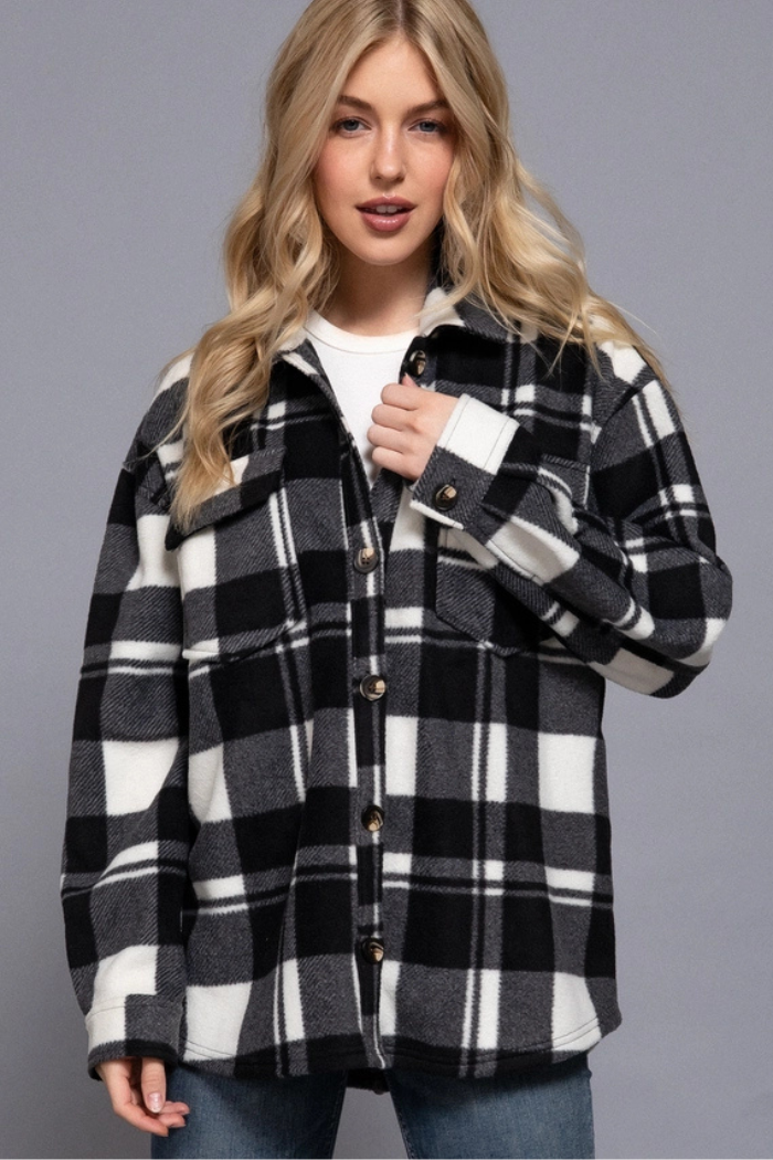 A young woman with blonde hair models a black and white checkered shacket over a white top. The shacket features a collared neckline, button-down front, and chest pockets with button flaps. 
