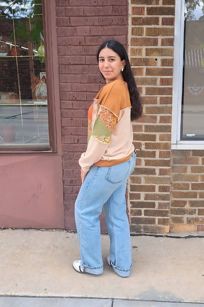 A young woman with long dark hair stands in profile, looking towards the camera. She is wearing a distinctive oversized top with patchwork elements, including sections of plaid and floral patterns in earthy tones, including rust, orange, mustard, and green, paired with blue jeans. The background is a brick wall.